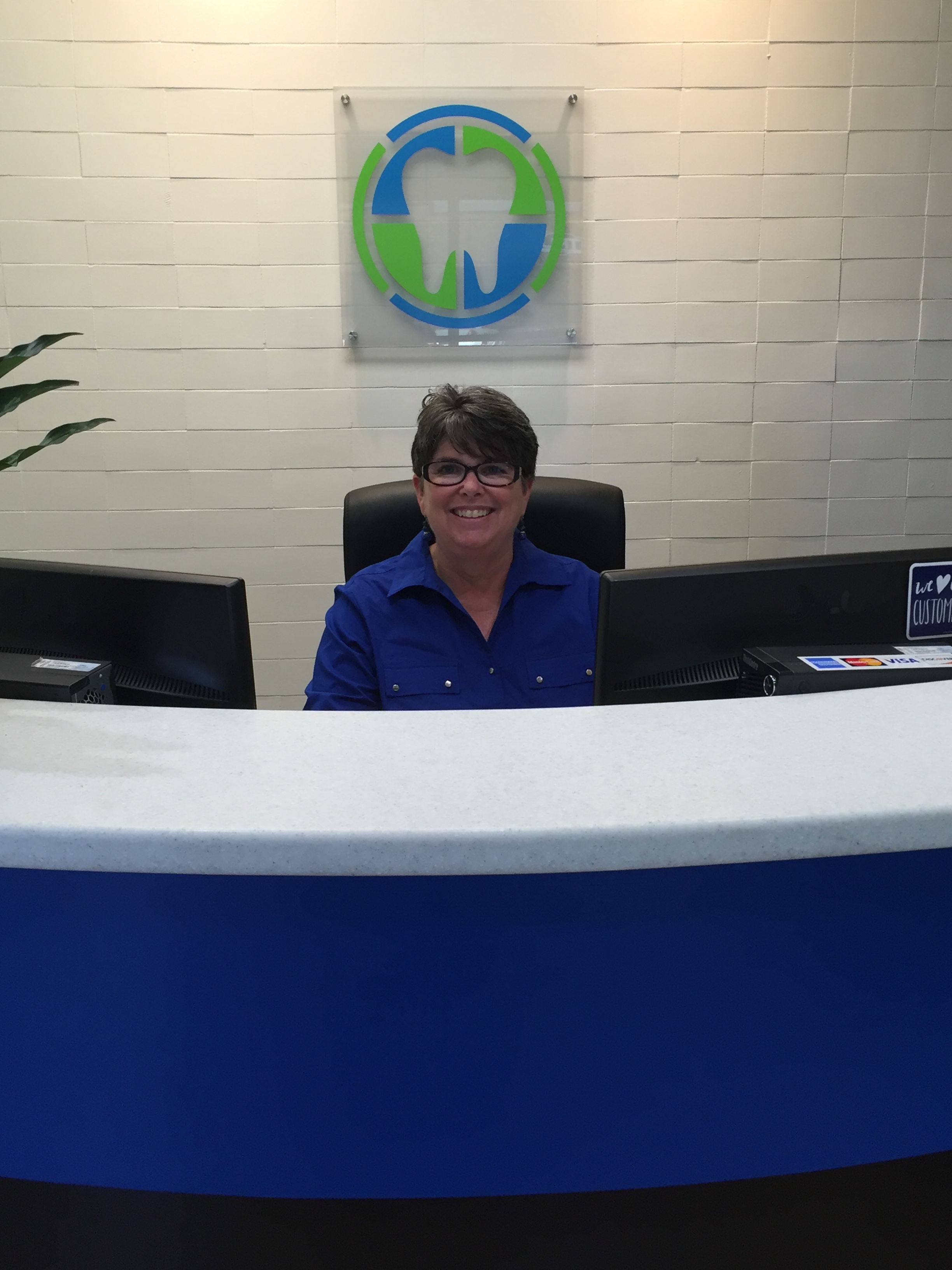Lynn sits behind a desk, smiling, in front of the Prime Family Dental logo.