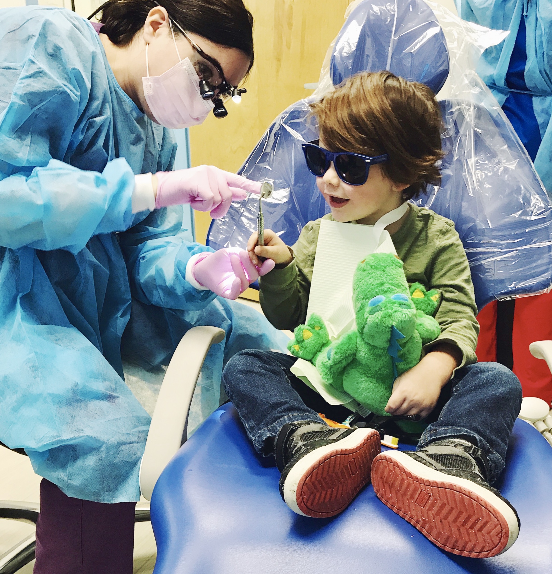 A young boy sits on a blue dental chair, wearing sunglasses and holding a stuffed alligator. He is smiling as Dr. Schtakleff shows him an instrument that she is using.